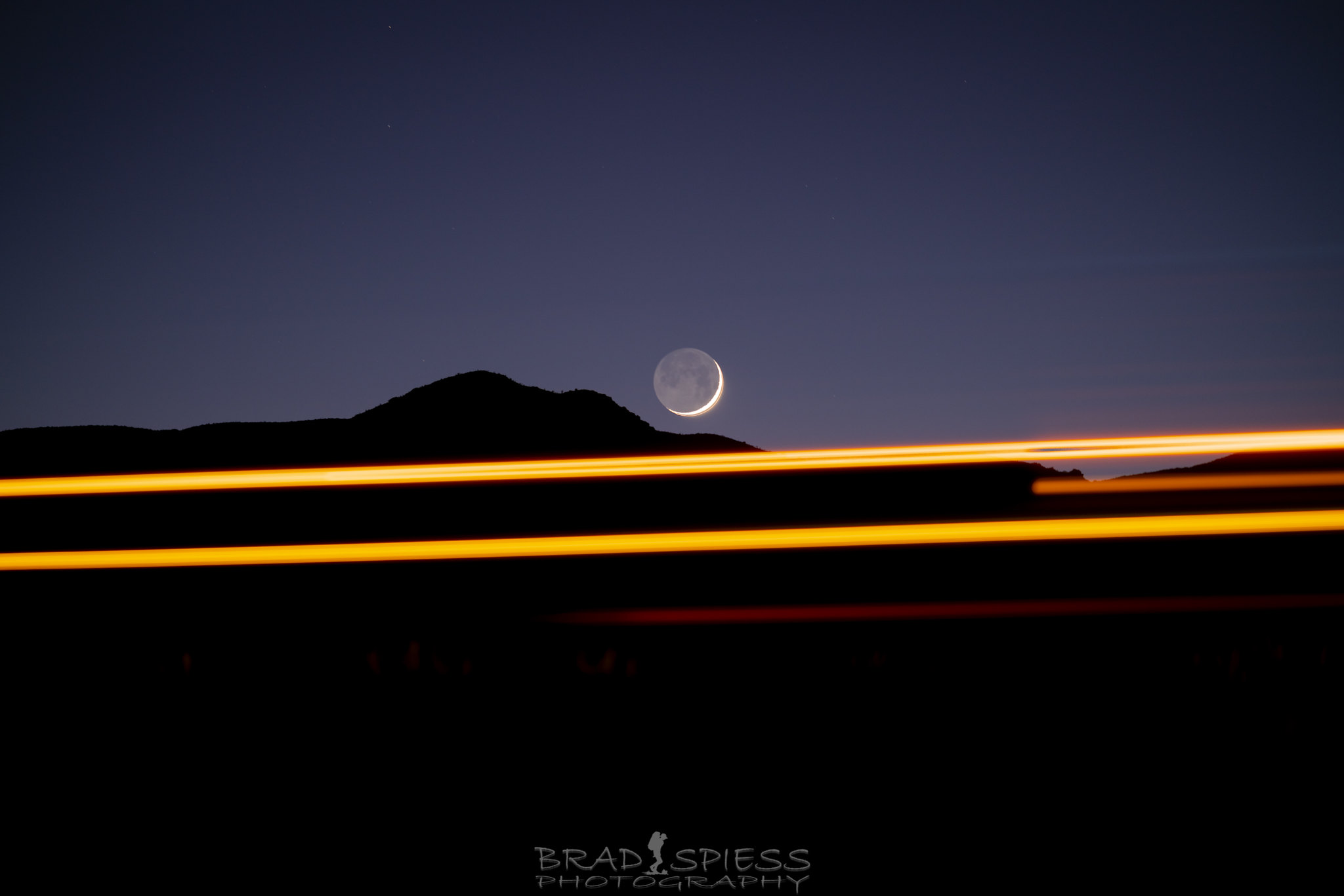 The descending crescent moon about to descend behind a mountain with the light trails from passing cars in the foreground.